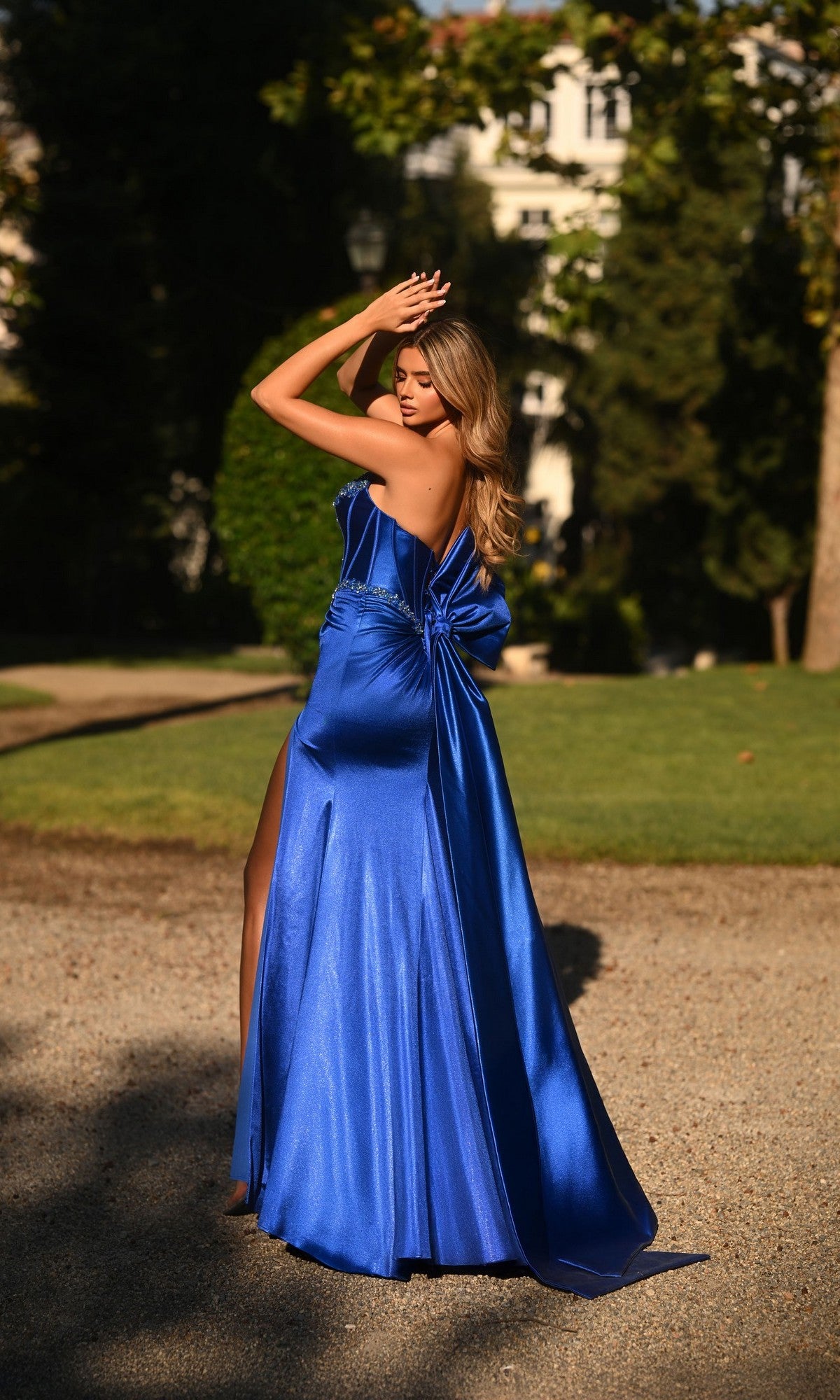 A woman outdoors on a gravel path poses with her arms raised, wearing the Alyce 62121 long formal dress—a shiny royal blue gown featuring a large bow on the back.